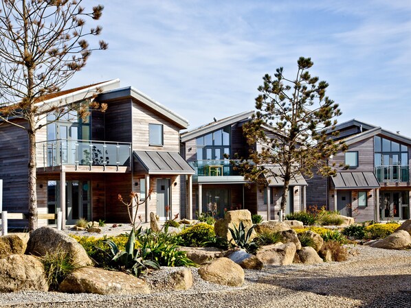 A modern multi-story house with a balcony, large windows, and a landscaped garden with rocks and plants.