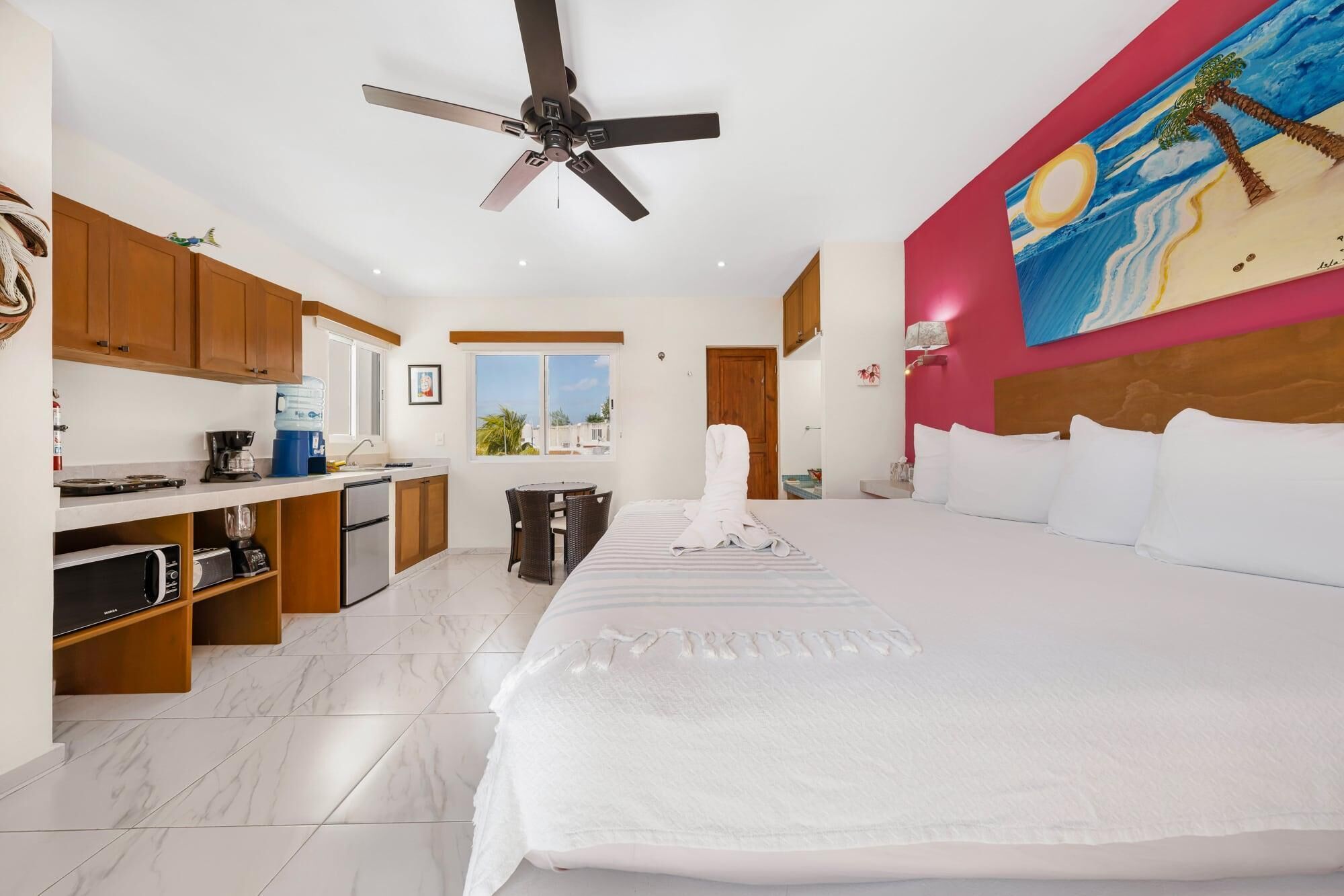Une chambre d’hôtel avec un lit, un ventilateur de plafond, un coin cuisine et un tableau au mur.