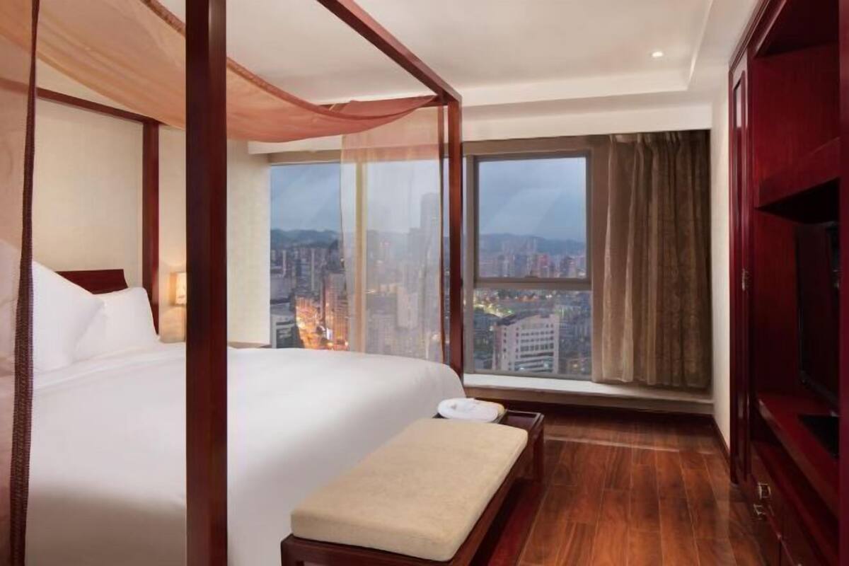 Een hotelkamer met een groot bed, een bankje, uitzicht op een stadsbeeld door een groot raam, en een kledingkast.