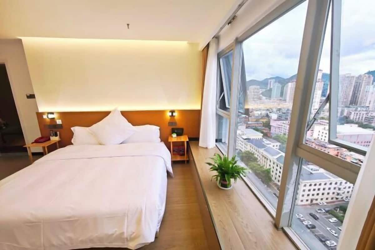 Hotelkamer met een groot bed, nachtkastjes, een kamerplant en stadszicht door grote ramen.