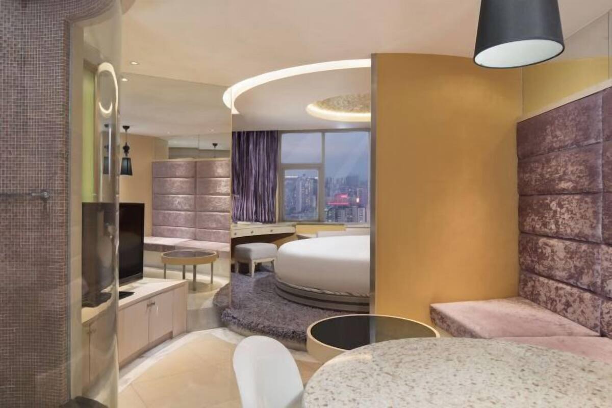 Een moderne hotelkamer met een gebogen bed, een televisie, een klein rond tafeltje en uitzicht op de stad door een groot raam.