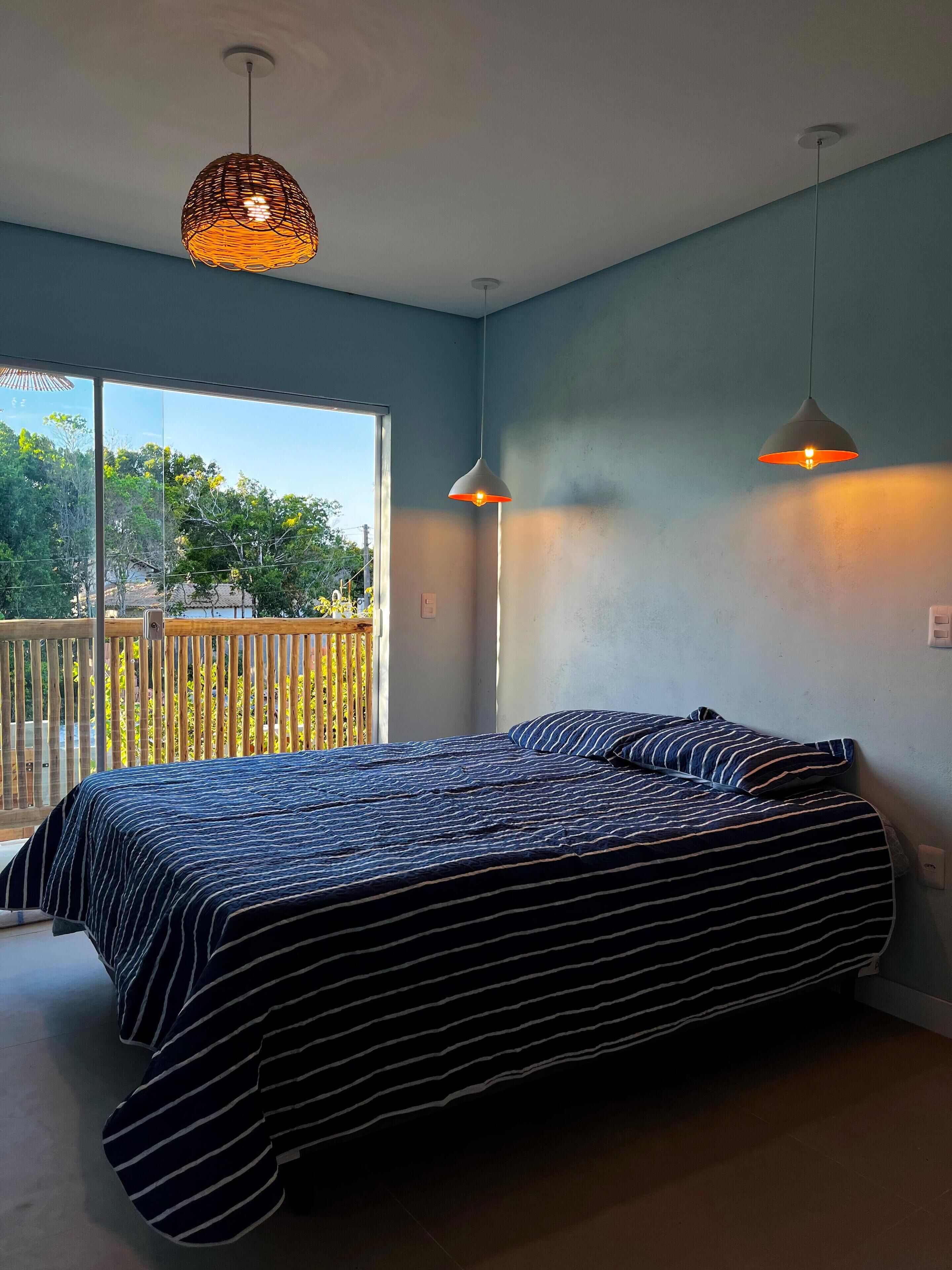 A bedroom with a large bed, two pendant lights, and a view of a balcony with a wooden fence.