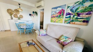 Ein Wohnzimmer mit einer Couch, einem Couchtisch und einem Essbereich mit Stühlen und einem Tisch. An den Wänden hängen zwei Gemälde.