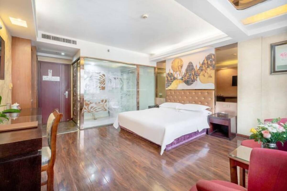 A hotel room with a bed, a desk, a chair, a nightstand, and a glass partition.