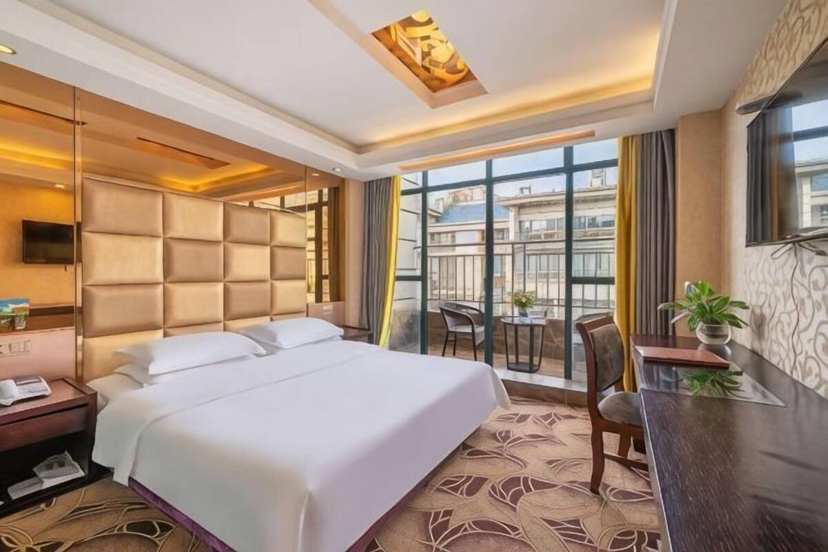 A hotel room with a large bed, a desk, a chair, a TV, and a balcony with a view of buildings.