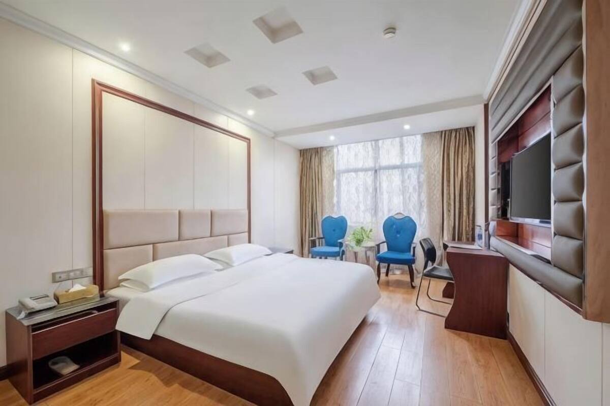A hotel room with a large bed, a TV, two chairs, a desk, and a window with curtains.