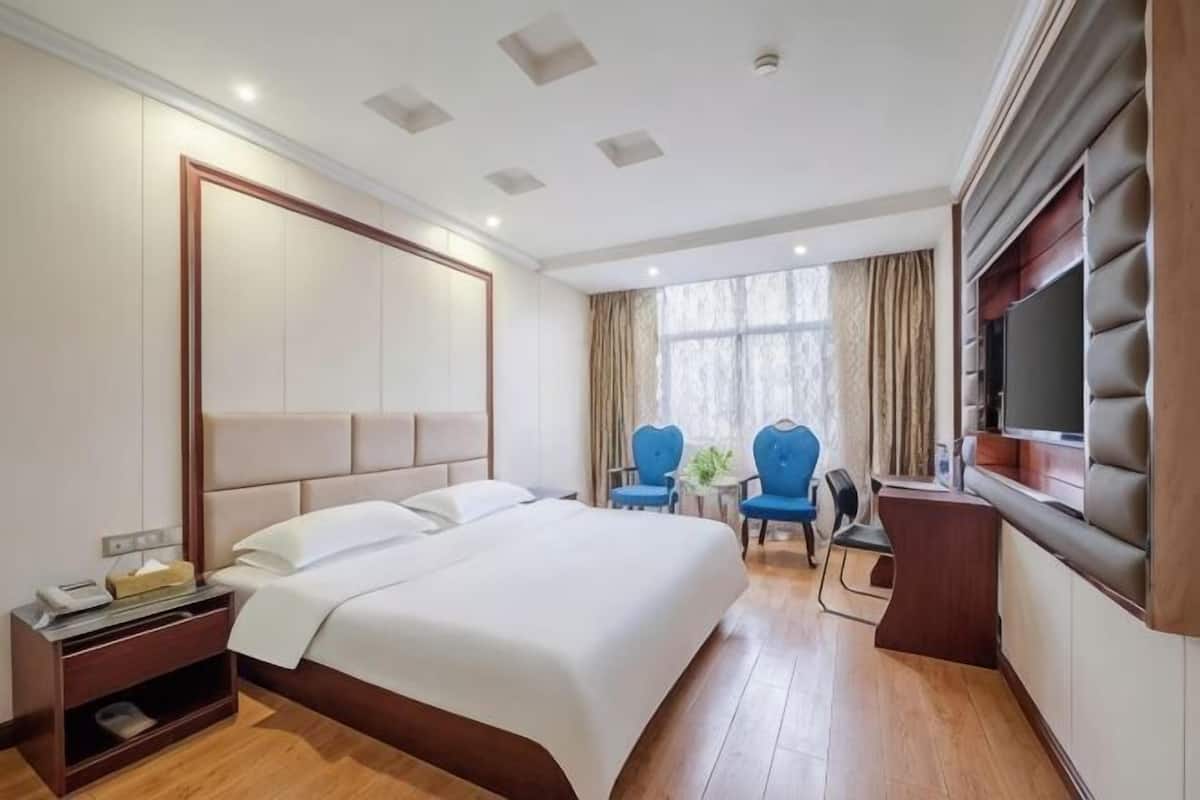 A hotel room with a large bed, a TV, two chairs, a desk, and a window with curtains.