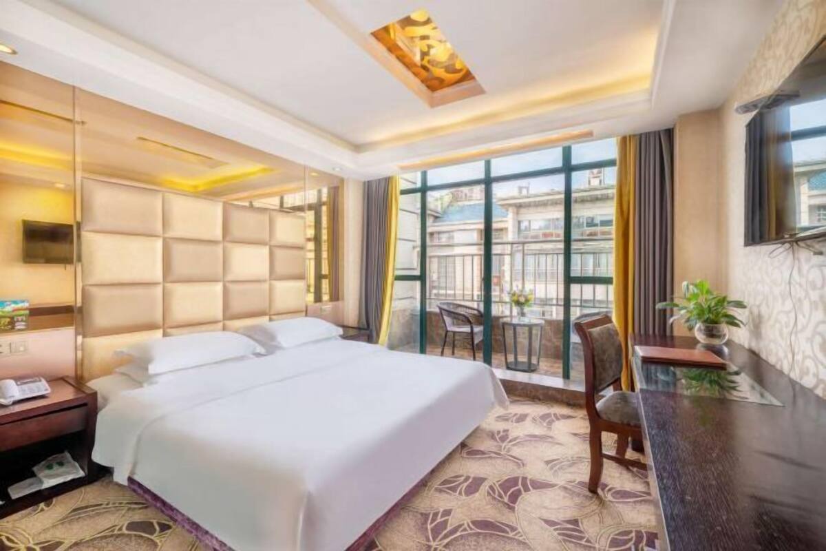 A hotel room with a large bed, a desk, a chair, and a balcony with a view.