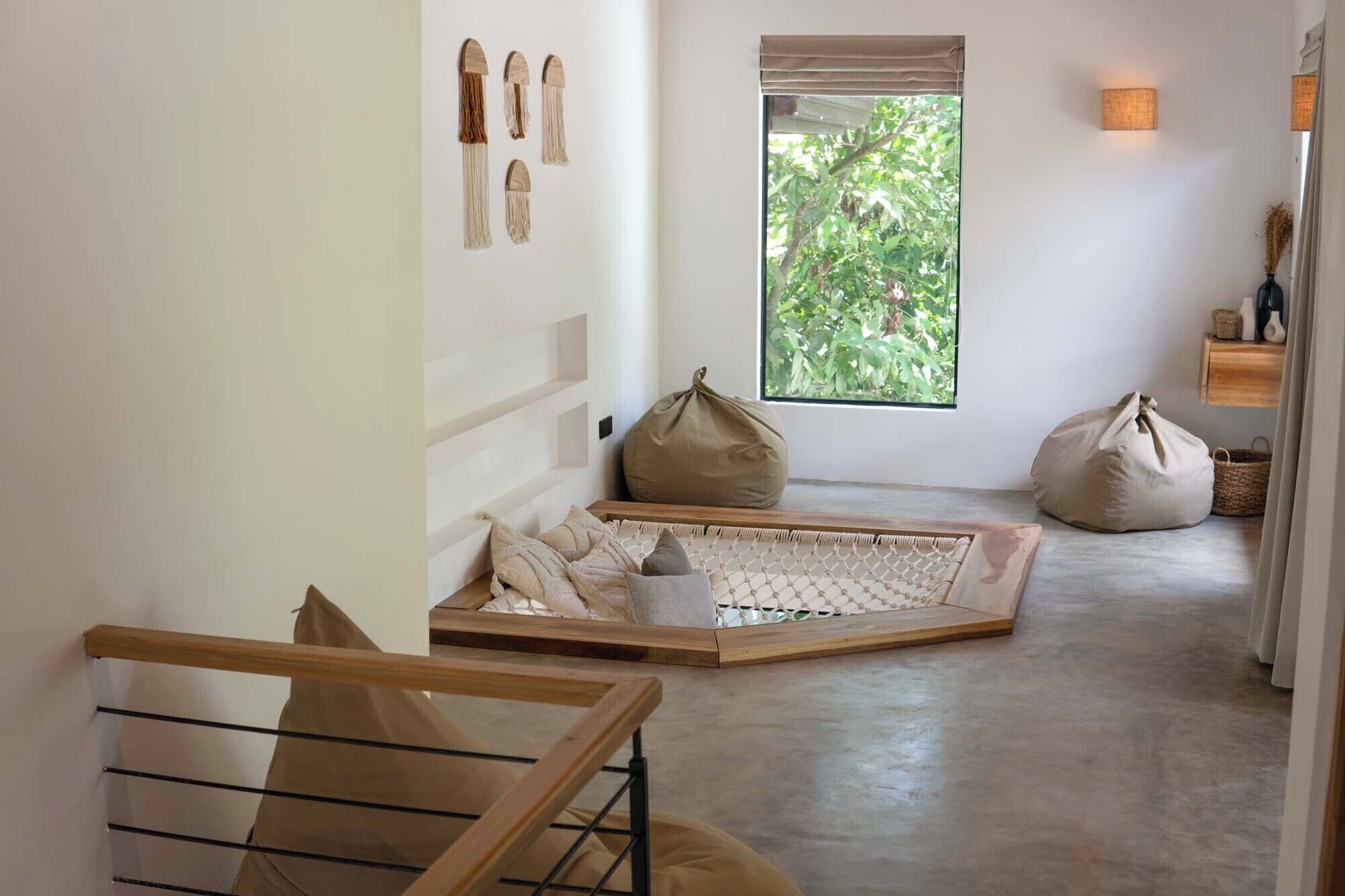 A minimalist interior with a hammock, bean bags, and a view of greenery through a window.