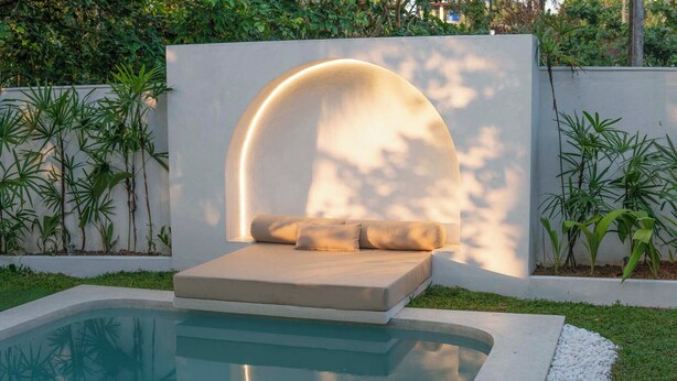 Un área de estar exterior moderna con piscina, rodeada de follaje y una pared blanca.