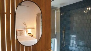 Habitación de hotel con un separador de madera, un espejo redondo, una cama con cabecera blanca, una mesita de noche con lámpara y vista a un área de ducha con pared de azulejos hexagonales.