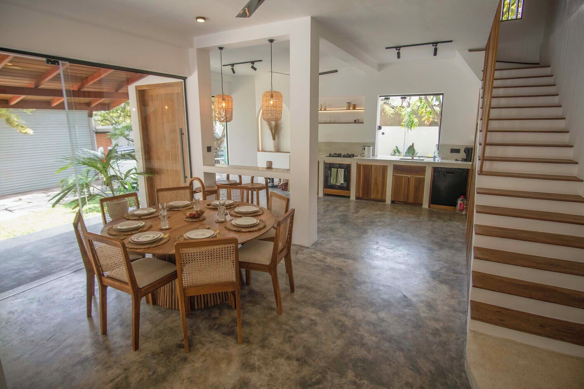 A spacious dining area with a round table set for six, surrounded by wicker chairs. A kitchen with modern appliances and a staircase leading to an upper level is visible in the background.