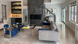 Una sala de estar moderna con un sofá gris, sillas azules, una mesa de centro y un televisor. Hay una escalera que conduce a un nivel superior.