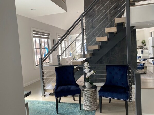 Una escalera moderna con acabado oscuro, dos sillones de terciopelo azul, una pequeña mesa redonda con una orquídea blanca y una vista de una zona de cocina al fondo.