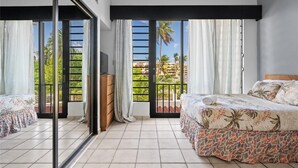 A bedroom with a bed, a dresser, and a TV. There are large windows with curtains and a view of palm trees and buildings outside.