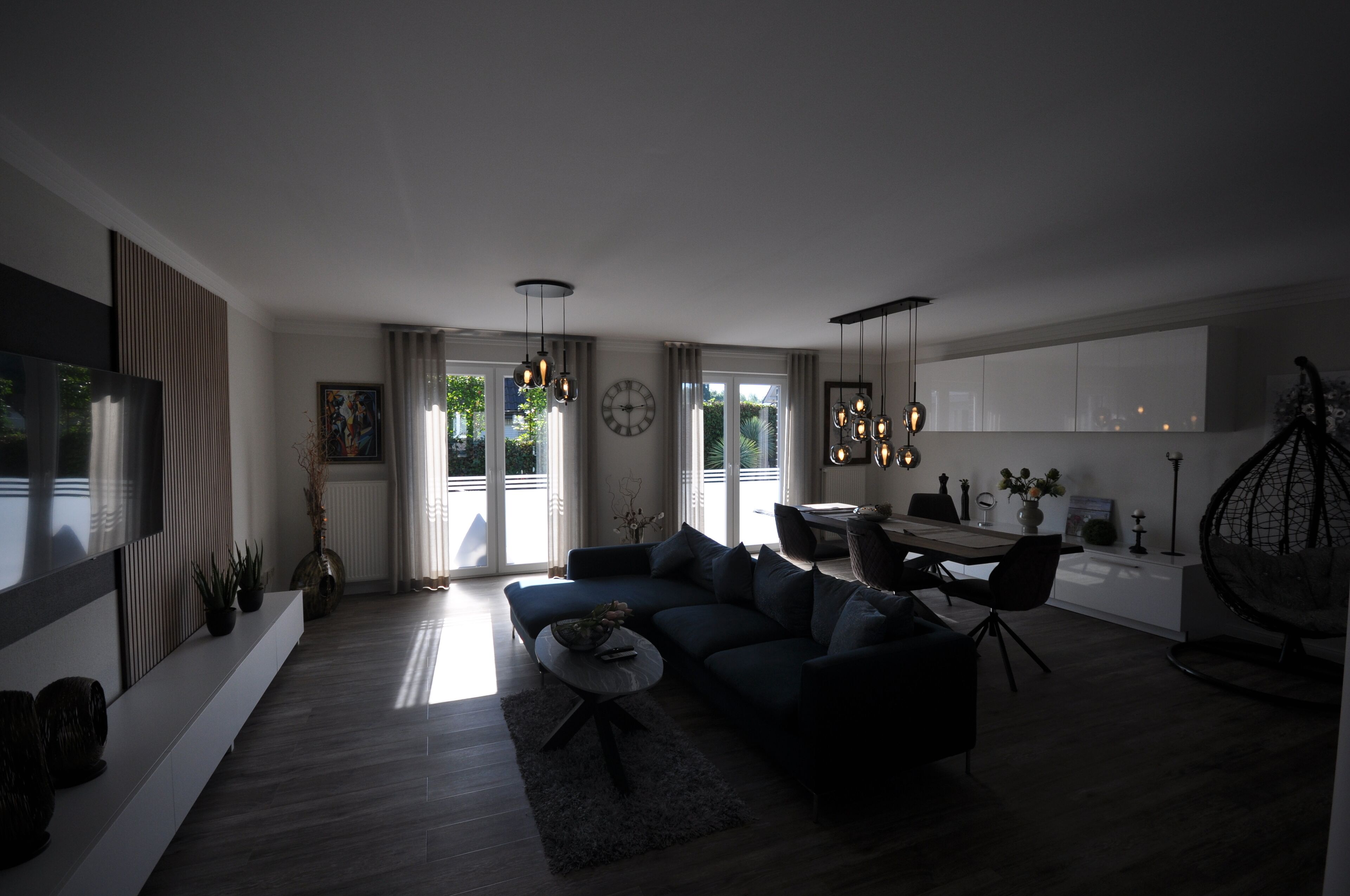 Ein modernes Wohnzimmer mit einer Couch, einem Couchtisch und einem Essbereich. Ein großes Fenster mit Vorhängen, einem Fernseher und einer Zierpflanze sind ebenfalls vorhanden.