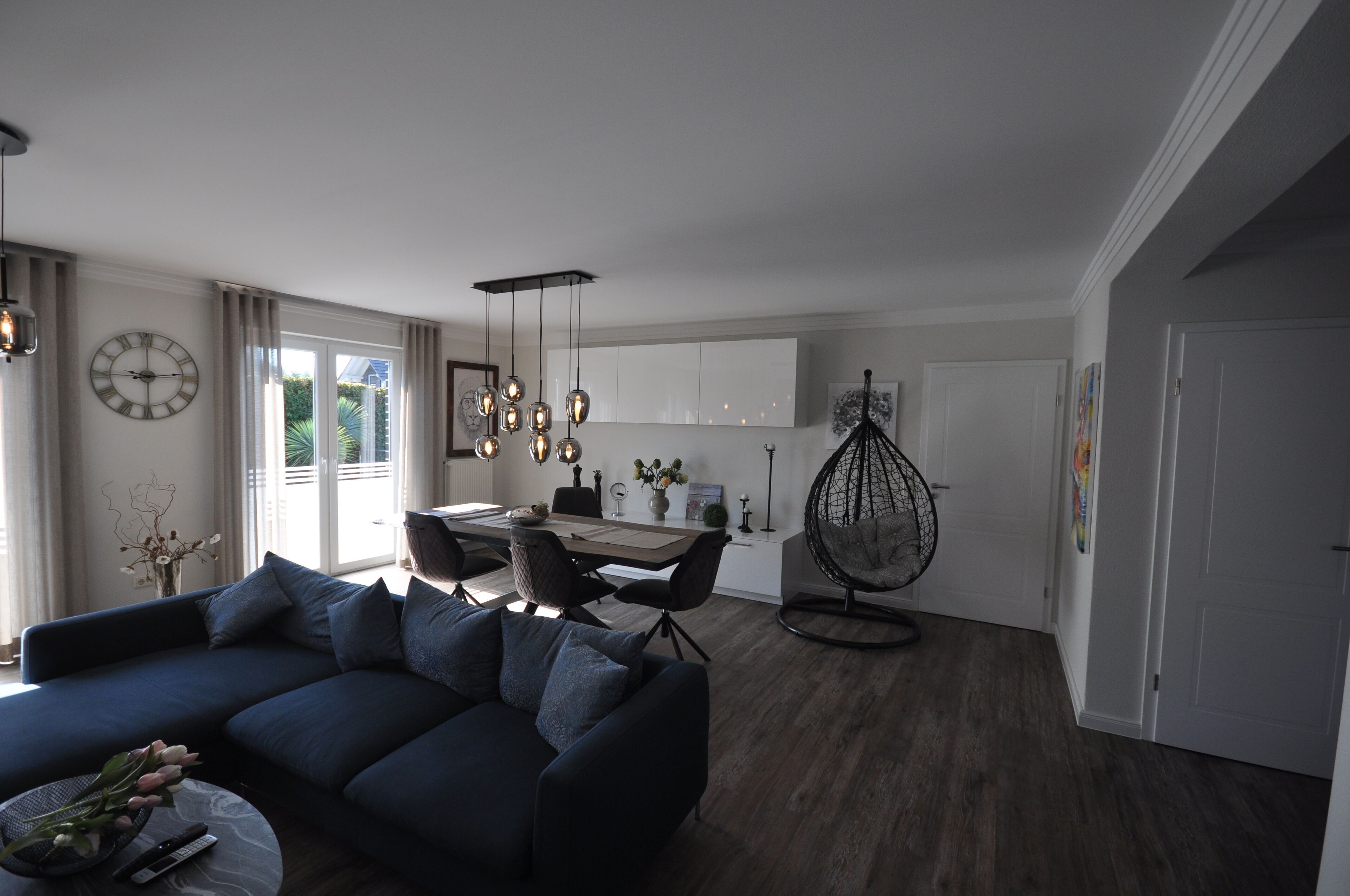 A modern living room with a dark blue sofa, a dining area with a table and chairs, and a hanging chair.