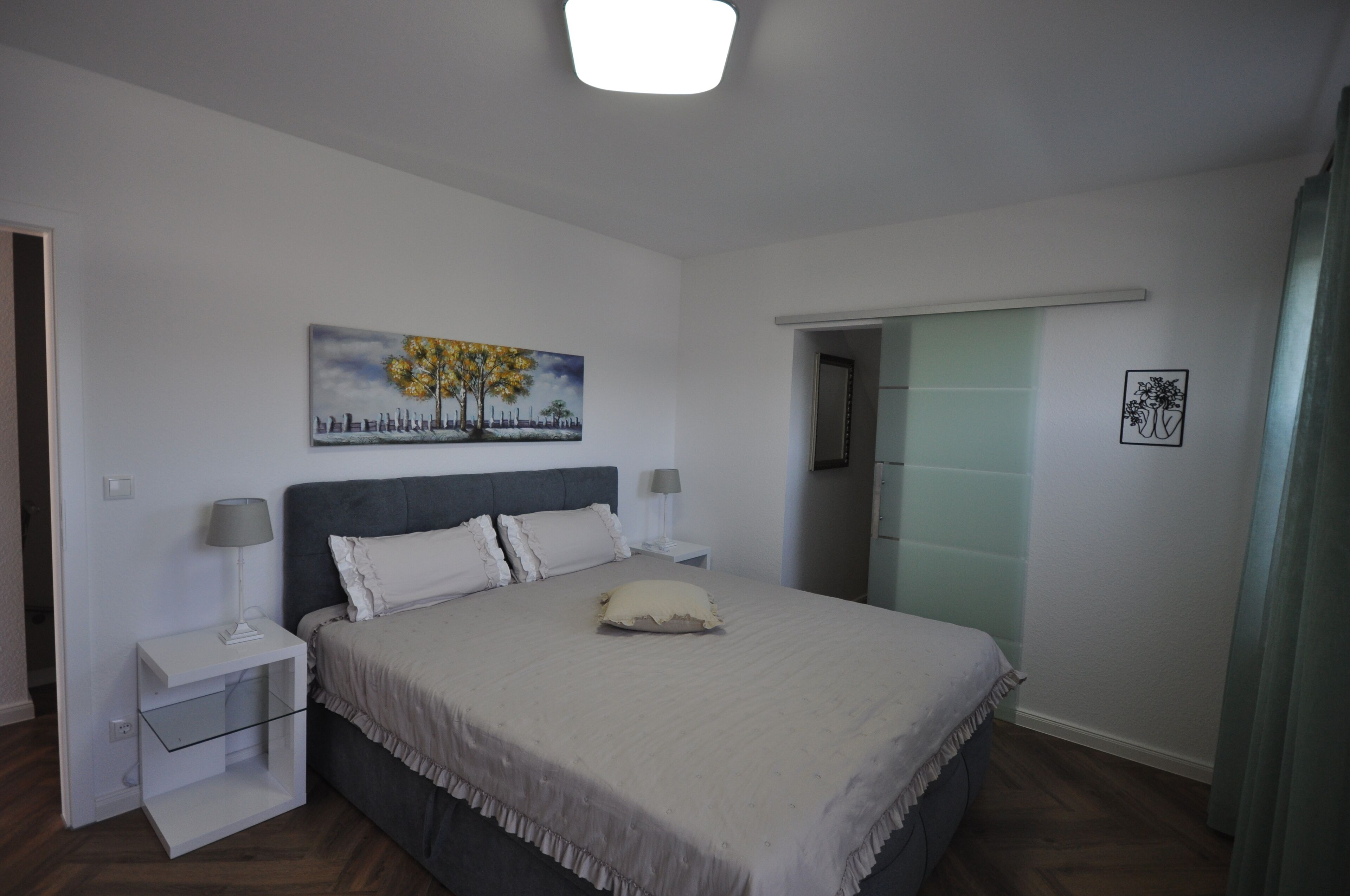 A bedroom with a bed, two bedside tables, a nightstand, a painting, and a door leading to another room.