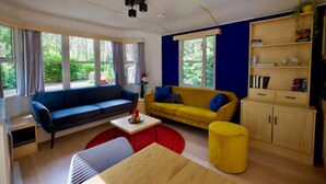 A modern living room with a blue sofa, yellow sofa, and a red ottoman. There is a wooden cabinet with books and a small plant.