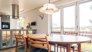A dining area with a round table and wooden chairs, a kitchen with a stainless steel range hood, and a large window with a view. - 4 Star Holiday Home in Pandrup (Pandrup)