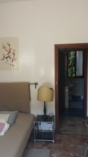 A room with a bed, a bedside table with a lamp, a painting on the wall, and a bathroom visible through an open door.