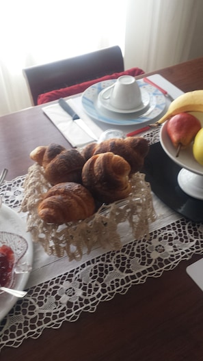 A table with a lace tablecloth, a plate of croissants, a fruit bowl with bananas and apples, and a cup and saucer.