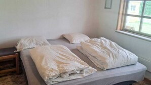 Un letto singolo con materasso grigio e lenzuola bianche, una panca in legno e una finestra con vista sul verde.