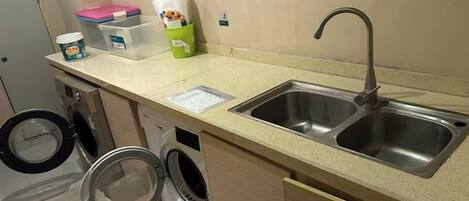 Una kitchenette con un lavabo, lavadora y una pequeña encimera con diversos objetos.