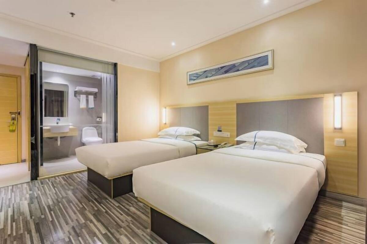 Quarto de hotel com duas camas, banheiro com porta de vidro e um quadro na parede.