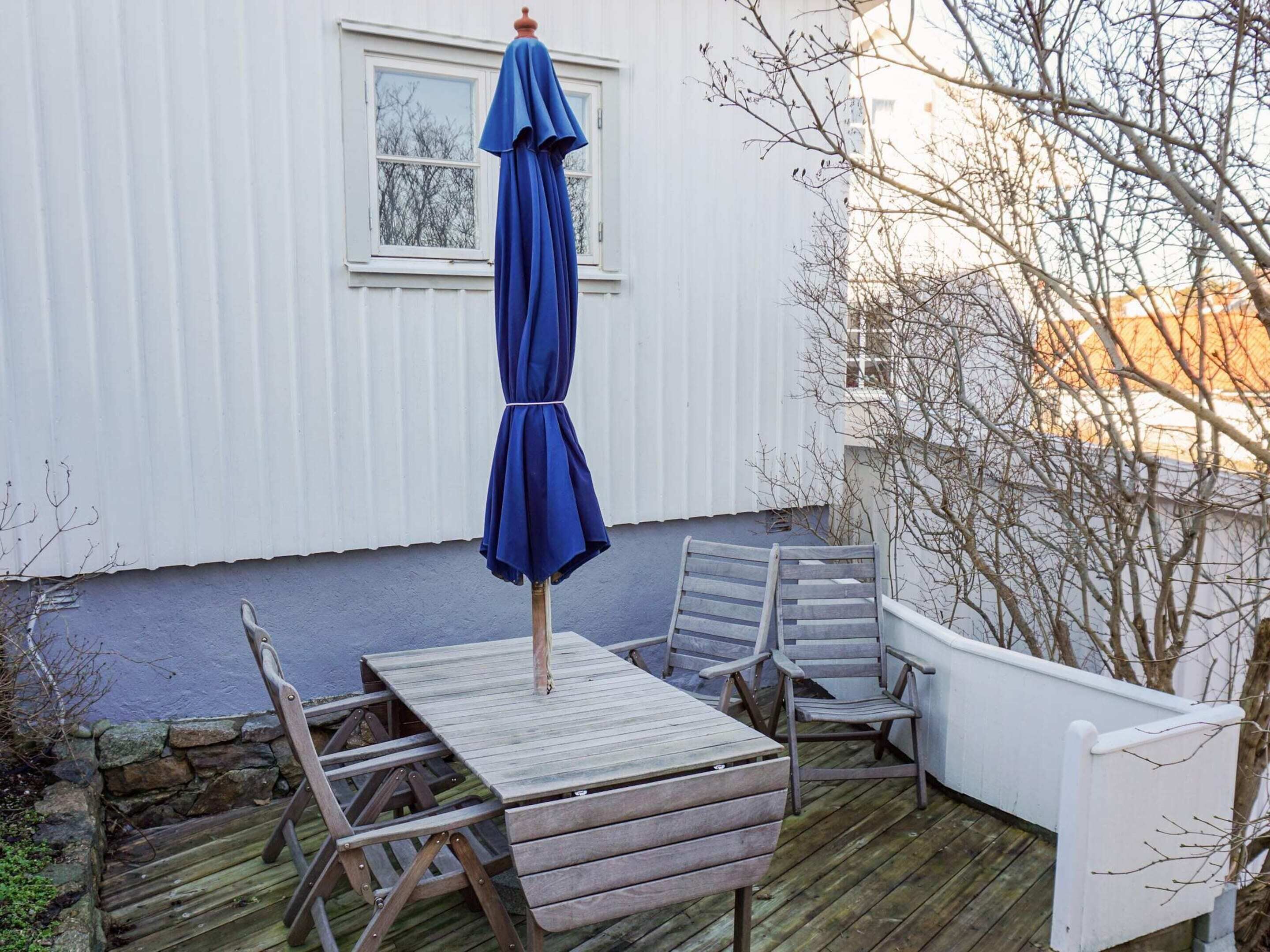 En träveranda med ett bord och stolar, ett blått parasoll och en vit byggnad i bakgrunden.