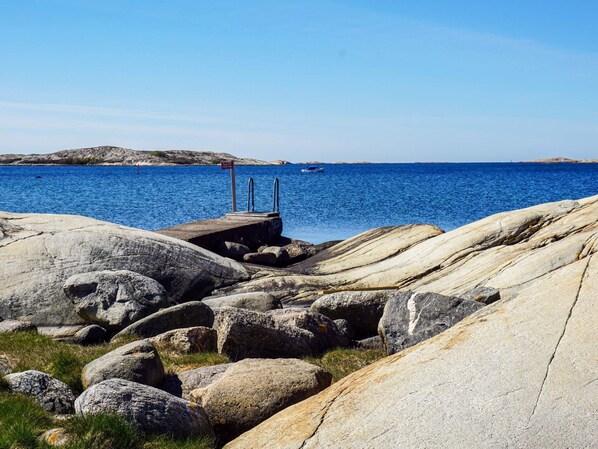 A rocky shoreline with a pier extending into the sea, a small boat anchored in the distance, and a clear blue sky.