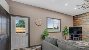 A living room with a gray sofa, a flat-screen TV on a wooden TV stand, a clock on the wall, and a door with a window. - Raccoon Lake Retreat (Rockville)