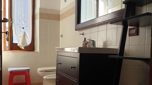 Une salle de bain avec des toilettes, un lavabo et un miroir. On y trouve un tabouret rouge et une fenêtre avec des rideaux.