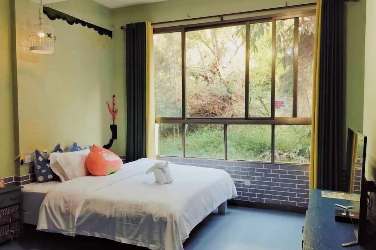 A bedroom with a bed, a window with a view of greenery, a desk with a computer, and a wall-mounted light fixture.