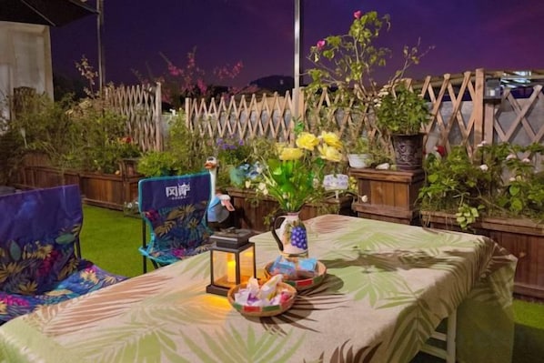 An outdoor dining area with a table set for two, surrounded by potted plants and a wooden fence.