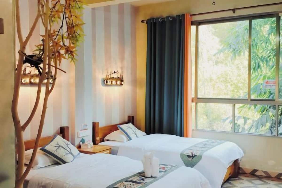 A hotel room with two beds, a large window with curtains, a wall-mounted light fixture, and a decorative tree branch.