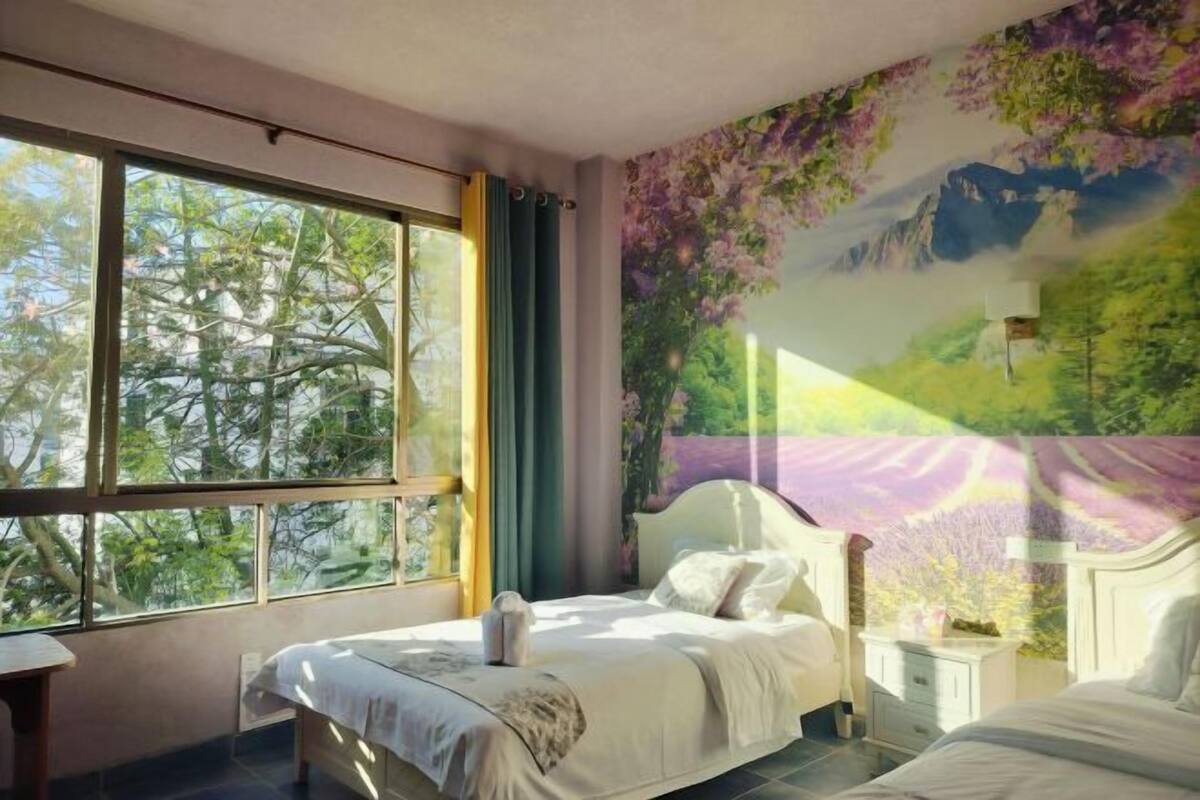 A bedroom with a large window, a bed, a nightstand, and a mural on the wall.