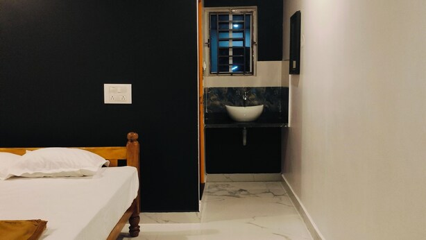 A bedroom with a wooden bed, a nightstand, a white wall, a window with blinds, and a bathroom with a sink and mirror. - Nature Valley Gokarna  (Kumta)