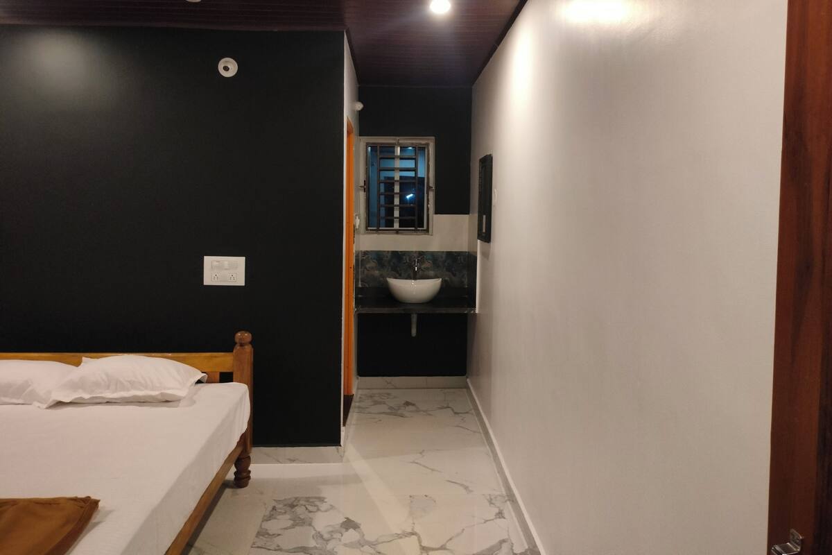A bedroom with a bed, a nightstand, a window, and a bathroom with a sink and mirror.