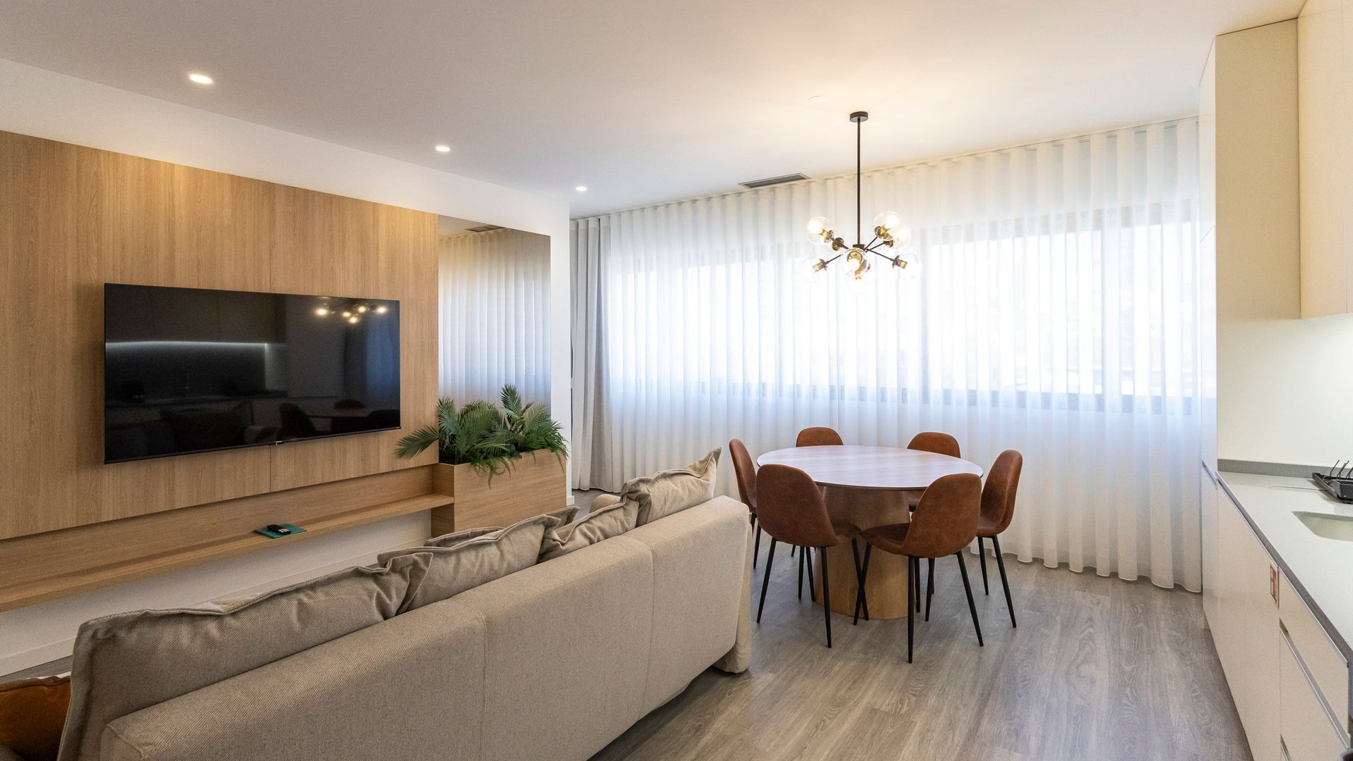 Una sala de estar moderna con un televisor de pantalla plana montado en la pared, un sofá color beige, una mesa de comedor con seis sillas y una araña.