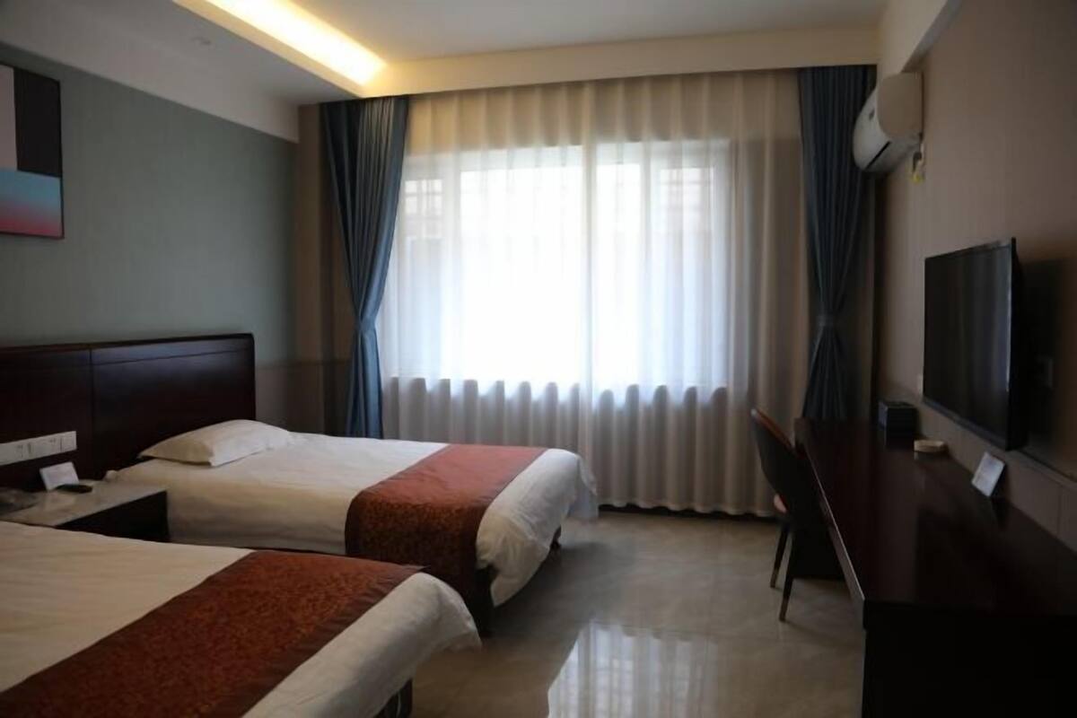 Quarto de hotel com duas camas, uma escrivaninha, televisão e uma janela grande com cortinas.