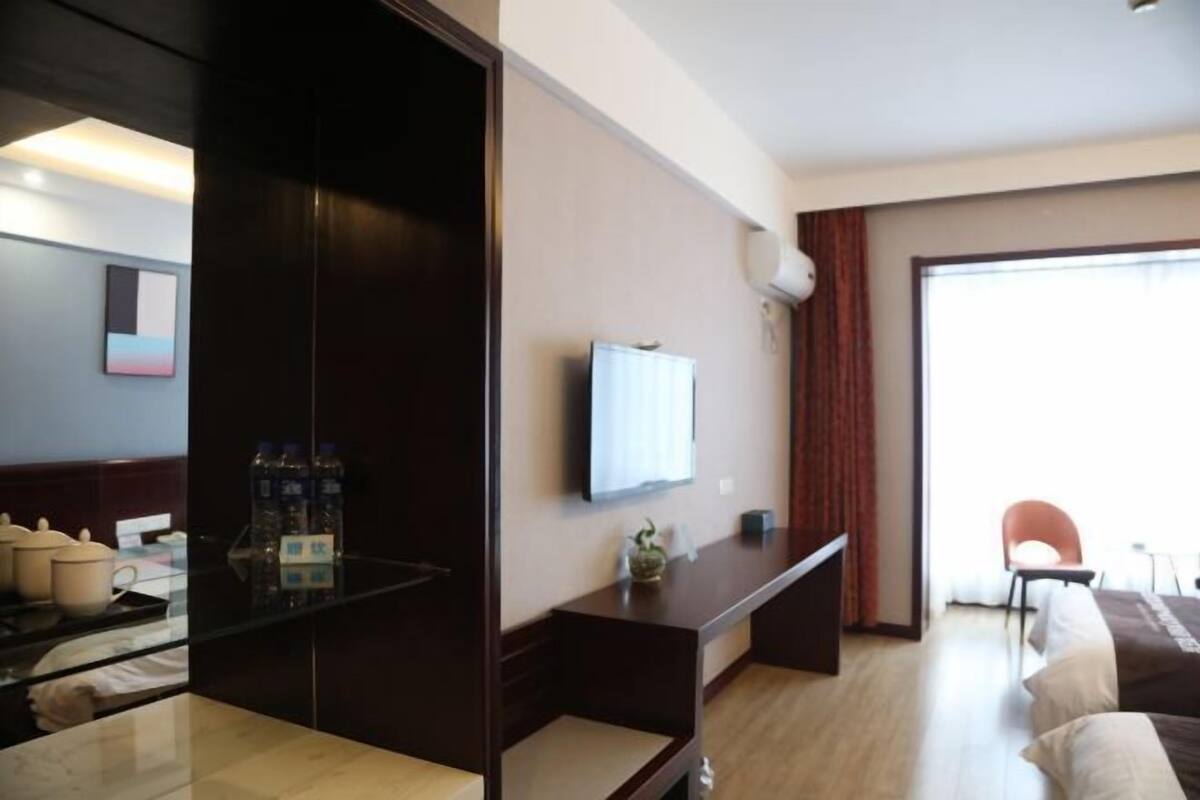 Quarto de hotel com televisão de tela plana, uma mesinha com uma planta, uma cadeira e uma cama com almofadas.