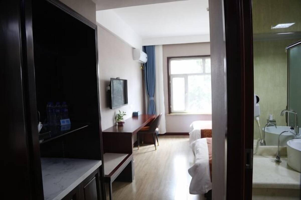 Quarto de hotel com cama, escrivaninha, cadeira, televisão, janela com cortinas e banheiro com pia e espelho.
