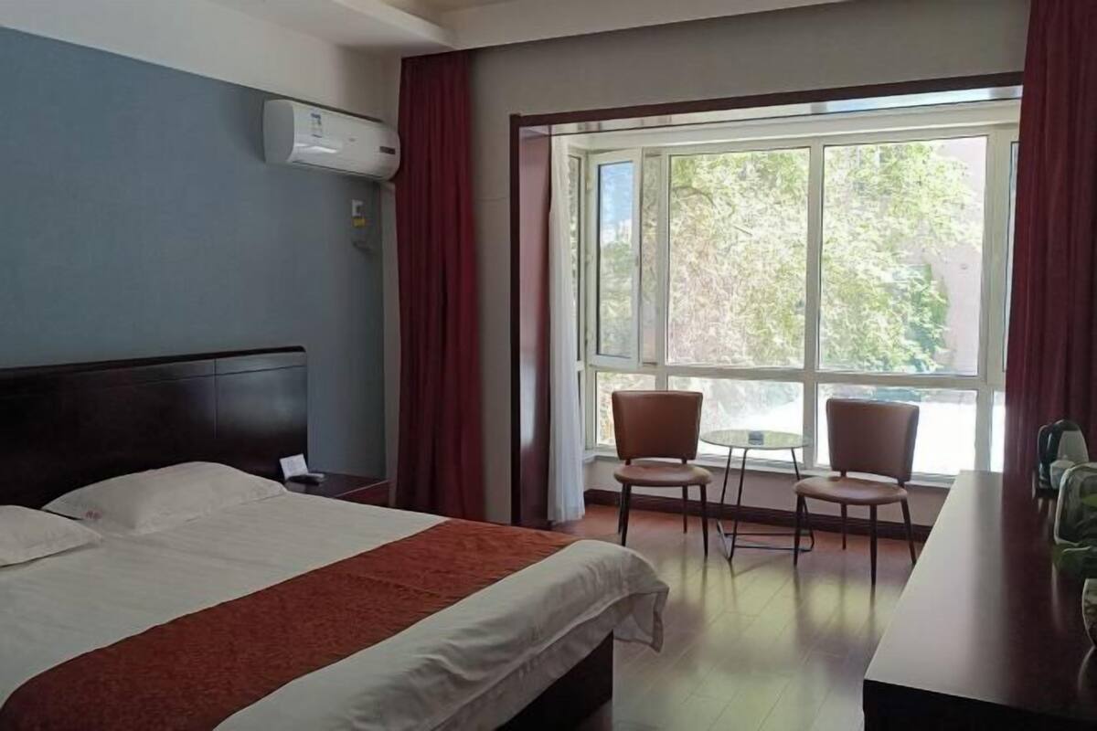 Quarto de hotel com cama, janela com cortinas, mesa e cadeiras, e um aparelho de ar condicionado.
