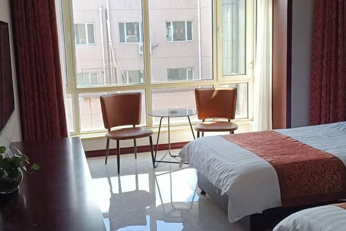 Quarto de hotel com cama, uma mesinha, duas cadeiras e uma janela grande.