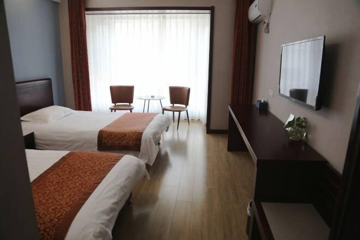 Quarto de hotel com duas camas, uma escrivaninha, uma cadeira, uma mesinha e uma televisão na parede.