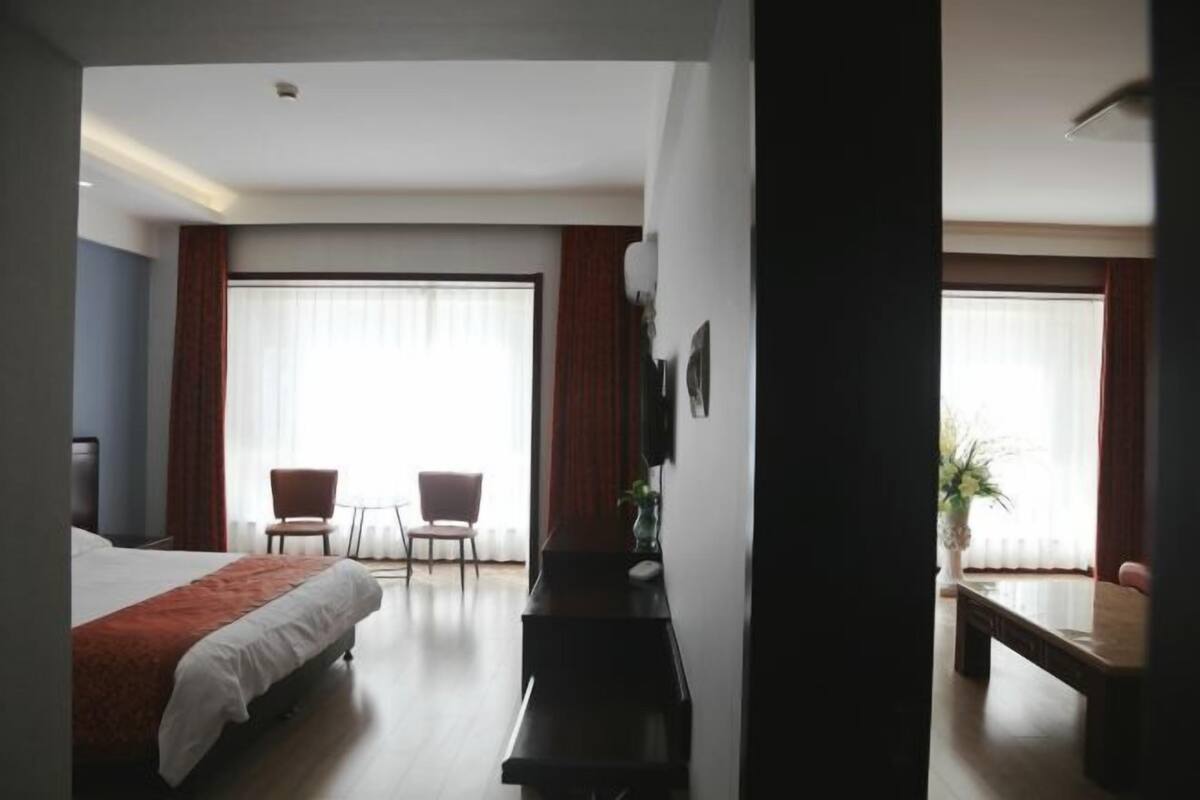 Quarto de hotel com cama, escrivaninha, duas cadeiras e uma janela grande com cortinas.