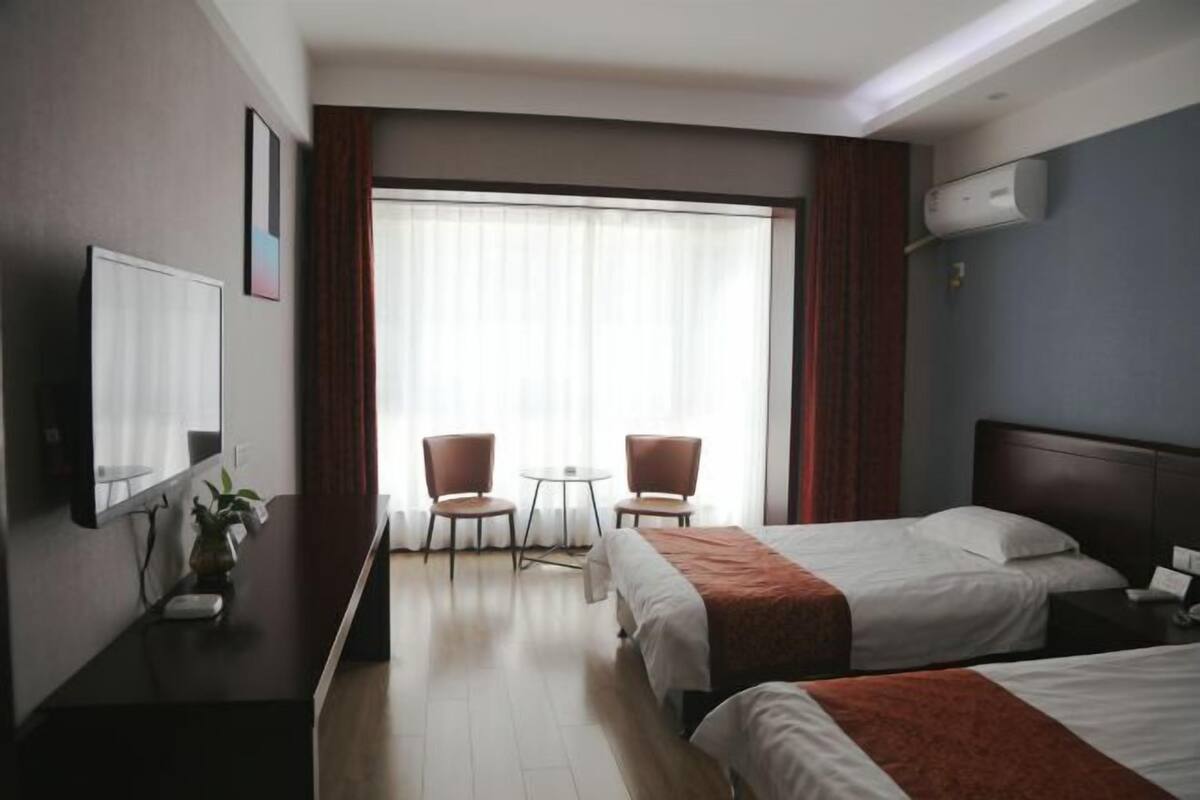 Quarto de hotel com duas camas, uma escrivaninha, uma cadeira, uma mesinha e uma janela com cortinas.