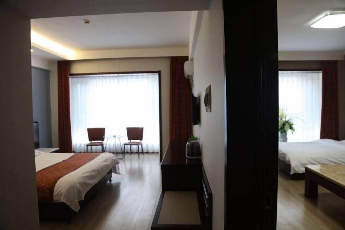 Quarto de hotel com duas camas, uma escrivaninha, duas cadeiras, uma televisão e uma janela grande com cortinas.