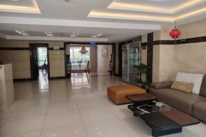 A hotel lobby with a reception desk, a seating area with a sofa and ottoman, a dining table, and a large window.